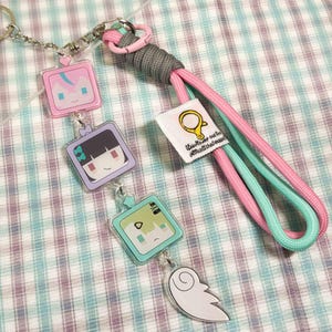 May include: A keychain featuring three square acrylic charms with cartoon faces, connected by silver metal rings to a pink and mint green paracord lanyard. The lanyard includes a grey wrapped section, a pink plastic ring, and a white fabric tag.
