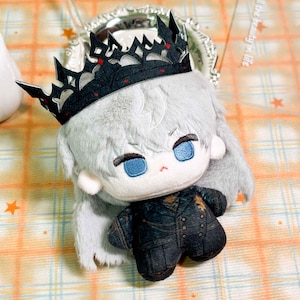 May include: A plush doll with light gray hair and blue embroidered eyes, wearing a black crown with red accents and a dark, textured double-breasted jacket. The doll features soft, fuzzy fabric and a small, simple facial expression with a tiny red mouth.