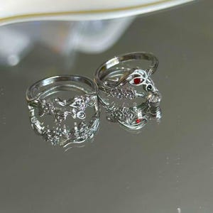 May include: Two silver rings with intricate designs. One ring features a snake design with red gemstone eyes. The other ring has a floral design. Both rings are displayed on a reflective surface.