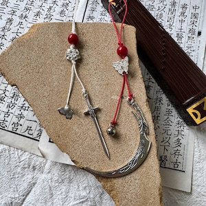May include: Two decorative hanging ornaments featuring silver-toned metal charms. One ornament has a white cord, a red bead, a flower charm, and a sword pendant. The other has a red cord, a red bead, a butterfly charm, a small bell, and a crescent moon.
