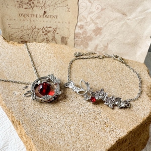 May include: A silver-toned necklace and bracelet set. The necklace features a circular pendant with a central red bead and leaf accents. The bracelet consists of a delicate chain with a floral and leaf motif, accented by a small red stone.