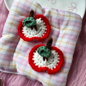 May include: Two handmade crochet apple slices featuring a bright red outer border, a white inner section, a small green leaf, and a brown stem. The apple slices are crafted with yarn and display a textured, looped stitch pattern throughout the design.