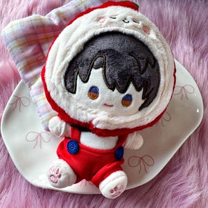 May include: A plush doll with dark hair wearing a white and red hooded cape with a small face embroidered on the hood. The doll has a white shirt, red overalls with blue buttons, and white feet with pink paw pad embroidery on the soles.