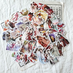 May include: A collection of assorted stickers featuring anime-style illustrations of characters in traditional East Asian clothing, with various red, white, and black colour schemes, layered over a piece of paper with printed Chinese characters.