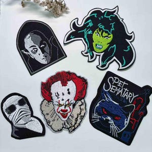 May include: A collection of embroidered patches depicting characters from horror films. The patches include a vampire, a green-skinned witch, a clown, a mummy, and a black cat. Each patch is black with detailed coloured stitching.