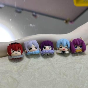 May include: Five small, colourful anime-style figurines. Each figurine has a unique hairstyle and colour, including red, lavender, purple, light blue, and dark purple. They are wearing scarves or collars in various colours.