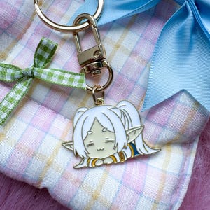May include: A gold-toned metal keyring featuring a white-haired anime character charm with pointed ears and a closed-eye expression. The charm is attached to a gold lobster clasp and ring, accented by a green gingham ribbon and a light blue satin bow.