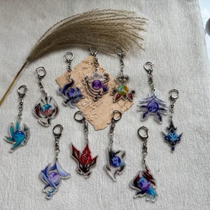 May include: A collection of eleven acrylic keychains featuring various colorful, stylized circular emblems and intricate geometric designs. Each charm is attached to a silver-toned metal lobster clasp and jump ring, displaying vibrant hues of blue, red, purple, and gold.