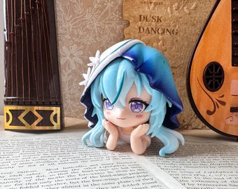 May include: A small anime-style figurine with light blue hair and purple eyes, wearing a white and blue hooded cloak with white flower accents. The character rests their chin on their hands, featuring a subtle smile and pink blush on their cheeks.