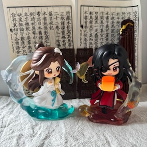 Puede incluir: Dos figuras estilo chibi con cabello oscuro. La figura de la izquierda viste túnicas blancas con detalles azules, rodeada de efectos de agua azul translúcida. La figura de la derecha viste túnicas rojas con un parche negro y sostiene una linterna naranja.