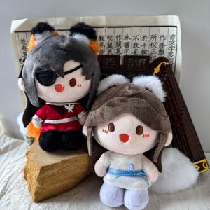 May include: Two plush dolls with embroidered facial features. The left doll has dark grey hair, orange fox ears, a black eye patch, and a red outfit. The right doll has brown hair, white ears, and a white outfit with a blue sash.