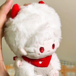 May include: A plush doll with white faux fur, featuring two small red horns on top of the head. The doll has embroidered red eyes, a small mouth, and a red fabric bandana around its neck. The plush material is soft and textured throughout.