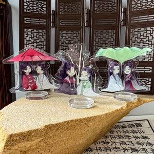 Könnte beinhalten: Drei durchsichtige Acryl-Aufsteller mit Anime-Figuren. Der erste zeigt zwei Figuren unter einem roten Regenschirm, der zweite zwei Figuren mit Bambus und der dritte zwei Figuren unter einem großen grünen Lotusblatt.
