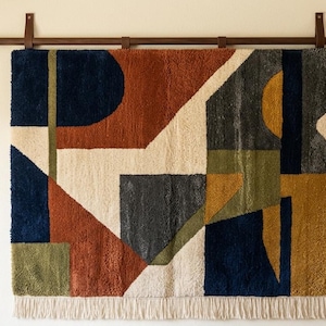 May include: Geometric wall hanging with a modern design. The textile features a mix of shapes in navy, rust, cream, gray, and olive green. It is mounted on a wooden rod with leather straps and has a fringed bottom.