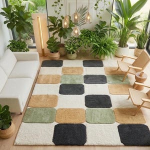 May include: A rectangular area rug featuring a grid pattern of rounded squares in shades of cream, charcoal gray, sage green, and tan. The rug is positioned on a light wood floor in a room with a white sofa, two wooden chairs, and many plants.