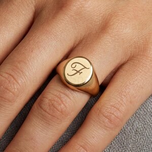 Puede incluir: Un anillo de sello de oro con una superficie lisa y pulida, grabado con la letra F en letra cursiva. El anillo presenta un diseño de parte superior redonda y plana con una banda sólida y un acabado metálico que refleja la luz.