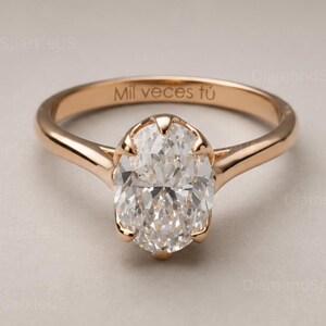 May include: A rose gold ring featuring a central oval-cut clear gemstone held by a multi-prong setting. The inner band is engraved with the text Mil veces tú in a clean, sans-serif font, showcasing a polished metal finish and a classic band design.