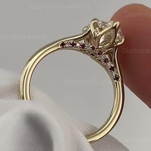 May include: A gold ring featuring a large, clear, round-cut center stone held in a six-prong setting. The band is accented with small, embedded red stones and tiny clear stones along the shoulders, creating a detailed, decorative metal design.