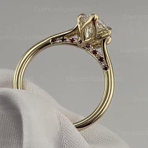 Puede incluir: Un anillo de oro con una gran piedra preciosa transparente central en un engaste de garras. La banda está decorada con pequeñas piedras rojas y transparentes alternadas incrustadas a lo largo de los hombros, creando un patrón decorativo.