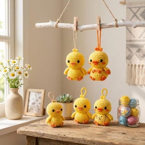 May include: Five small, round, yellow crocheted chick ornaments with orange beaks and feet. Two chicks hang from a white wooden branch by twine, while three sit on a wooden surface. Each chick has black bead eyes and a textured yarn finish.