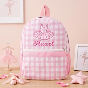 May include: A pink and white gingham backpack featuring an embroidered ballet tutu and the name Hazel in pink script. The backpack includes a front zippered pocket, a top handle, and side mesh pockets, constructed with a textured fabric finish.