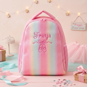 May include: A pink backpack with a vertical rainbow gradient and glittery finish. The front features embroidered text reading Freya above a pair of ballet slippers. It has a top handle, dual zipper closure, and side mesh pockets for storage.