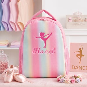 May include: A pink, blue, and white ombre backpack featuring an embroidered silhouette of a dancer and the name Hazel in cursive script. The backpack has a top handle, a zippered main compartment, and side mesh pockets for storage and organization.