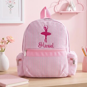 May include: A light pink and white seersucker backpack featuring an embroidered dark pink ballerina silhouette and the name Hazel in cursive script. The bag includes a main zippered compartment, a front zippered pocket, and two side pockets.