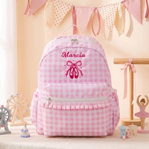 May include: A light pink and white gingham backpack featuring a ruffled front pocket and a top handle. The front is embroidered with the name Marcia in dark pink script above a graphic of dark pink ballet slippers with tied ribbons.