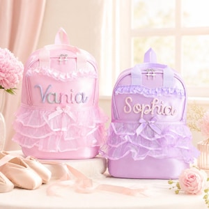 May include: Two backpacks in light pink and lavender, each featuring a ruffled tulle overlay, a central bow, and a name embroidered in cursive script. The pink backpack displays the name Vania, while the lavender backpack displays the name Sophia.