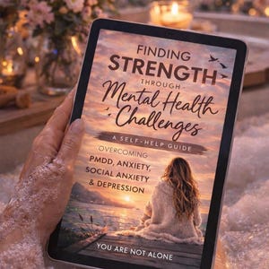 May include: A tablet displaying the cover of a self-help guide titled "Finding Strength Through Mental Health Challenges." The cover features a woman sitting on a dock at sunset. The guide addresses PMDD, anxiety, social anxiety, and depression.
