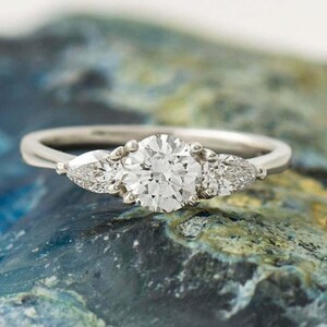 May include: A silver-toned ring featuring a large, round, brilliant-cut center stone flanked by two smaller, pear-shaped stones on either side. The band is smooth and metallic, showcasing a classic three-stone design with clear, faceted gems.