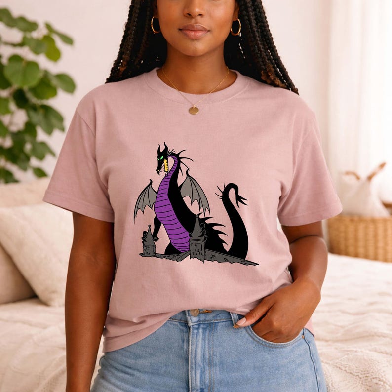 May include: A light pink t-shirt featuring a cartoon dragon design. The dragon is black with purple accents and gray wings. The dragon is sitting on a stone structure. The shirt is made of a soft material.