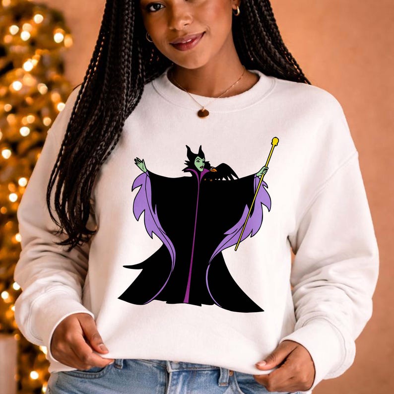 May include: White sweatshirt featuring a cartoon illustration of a character in a black and purple cloak, holding a golden staff. The character has green skin and a black headdress. A black bird is perched on the character's shoulder.