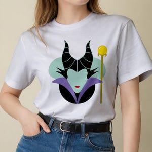 May include: White t-shirt featuring a stylized illustration of a character with black horns, a green face, and a purple collar. A gold staff with a yellow orb is to the right of the character.