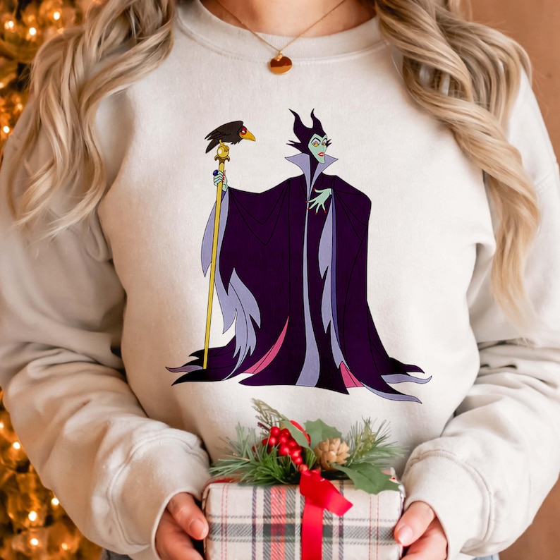 May include: A light beige sweatshirt featuring a graphic of a cartoon villain in a long, dark purple cloak, holding a staff with a raven on top. The character has green skin and a stern expression.