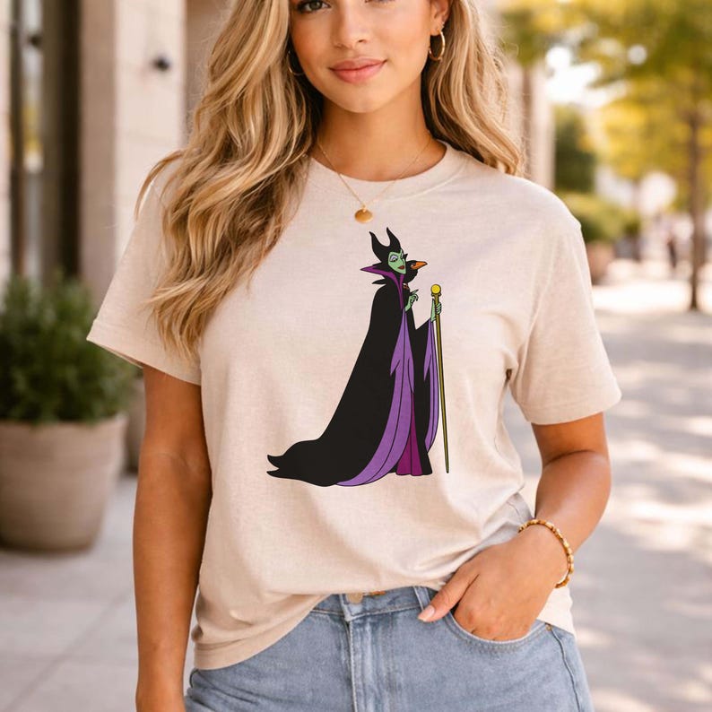 May include: Beige t-shirt featuring a cartoon illustration of a character in a black and purple cloak, holding a staff. The character has green skin and a horned headdress. The shirt is worn by a person in blue jeans.