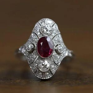 Peut inclure: Une bague argentée ornée d'un rubis rouge foncé taille ovale en son centre. La monture allongée est incrustée de nombreux petits diamants blancs ronds disposés en motif géométrique, avec trois diamants ronds plus grands placés en haut, en bas et sur les côtés.