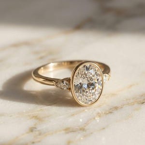 May include: A gold engagement ring with an oval-cut diamond center stone. The ring features a bezel setting and two smaller accent stones on each side. The band is a simple, polished gold. The ring is displayed on a marble surface.