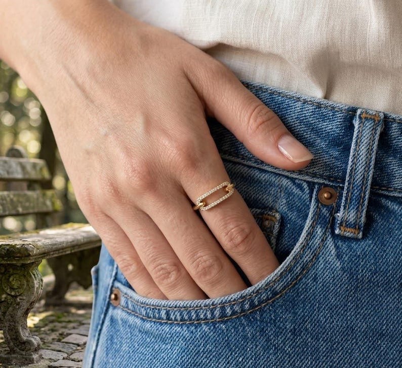 Pode incluir: Um anel dourado com um design retangular, incrustado com pequenas pedras preciosas transparentes. O anel &eacute; usado em um dedo, com a m&atilde;o repousando em um bolso de jeans azul. O fundo est&aacute; desfocado, sugerindo um ambiente ao ar livre.