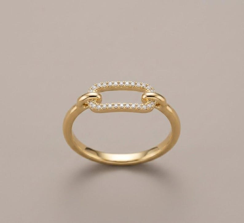 Pode incluir: Um anel dourado com um design retangular em forma de elo. O centro retangular &eacute; incrustado com pequenas pedras preciosas transparentes. O anel tem um acabamento liso e polido sobre um fundo neutro.