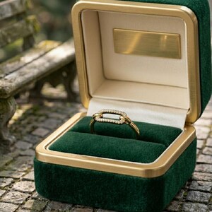 Pode incluir: Um anel de ouro com um design retangular, incrustado com pequenas pedras transparentes, repousa em uma caixa de anel de veludo verde. A caixa tem uma borda dourada e uma placa dourada dentro da tampa. A caixa de anel est&aacute; aberta.
