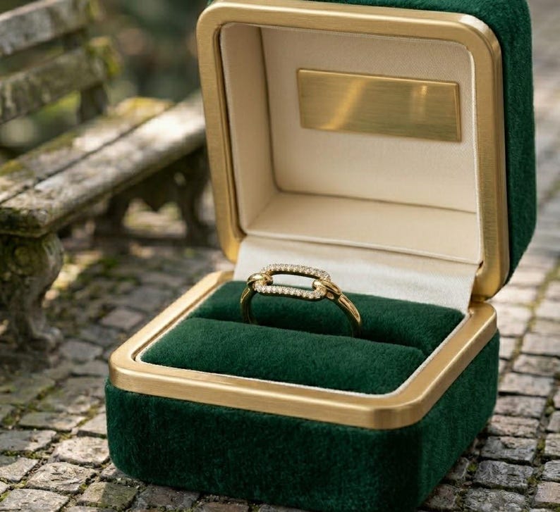 Pode incluir: Um anel de ouro com um design retangular, incrustado com pequenas pedras transparentes, repousa em uma caixa de anel de veludo verde. A caixa tem uma borda dourada e uma placa dourada dentro da tampa. A caixa de anel est&aacute; aberta.