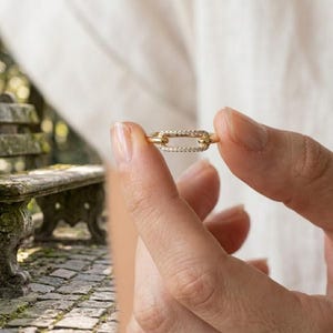 Pode incluir: Um anel dourado com um design de elo retangular, incrustado com pequenas pedras preciosas transparentes. O anel &eacute; segurado pelos dedos de uma pessoa, mostrando seus detalhes delicados. O design do anel lembra um elo de corrente. O fundo est&aacute; desfocado.