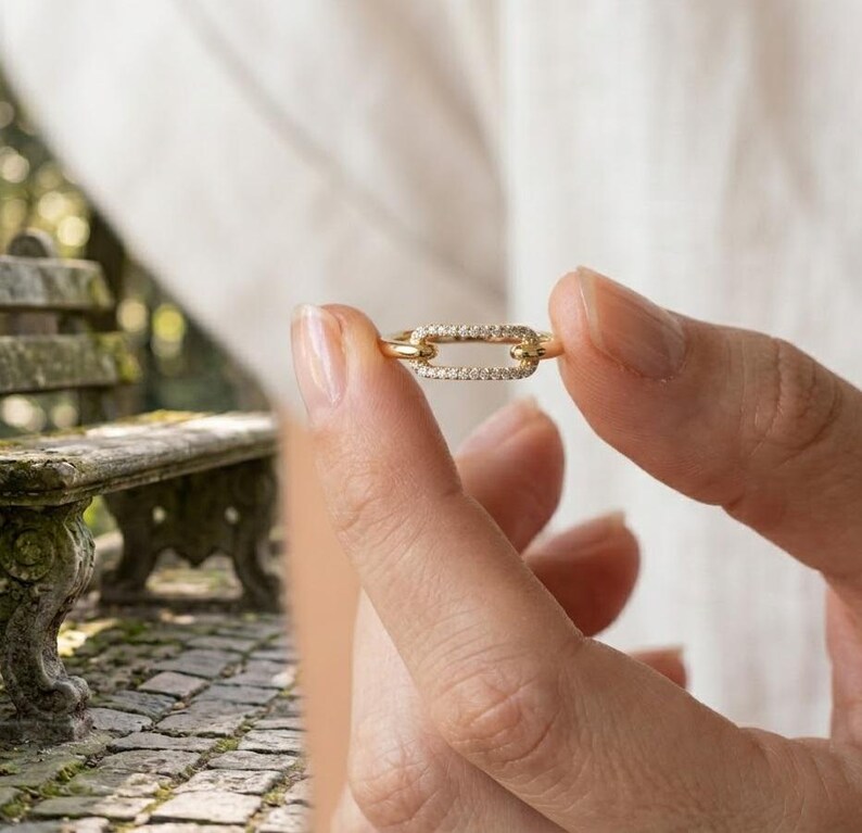 Pode incluir: Um anel dourado com um design de elo retangular, incrustado com pequenas pedras preciosas transparentes. O anel &eacute; segurado pelos dedos de uma pessoa, mostrando seus detalhes delicados. O design do anel lembra um elo de corrente. O fundo est&aacute; desfocado.