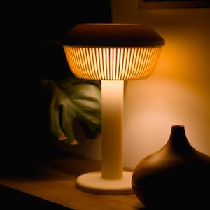 Puede incluir: Una lámpara de mesa moderna con una luz cálida. La lámpara tiene una base cilíndrica y una pantalla con listones verticales, coronada por una tapa marrón oscuro. Un jarrón de madera está cerca. La lámpara es una adición elegante a cualquier decoración.