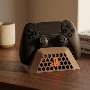 Puede incluir: Un mando negro de PlayStation 5 descansa sobre un soporte marrón con un diseño de panal y un logotipo naranja de PlayStation. El mando tiene botones negros y una luz azul. El soporte está sobre una superficie de madera.