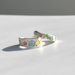 May include: Two silver-toned metal rings featuring colorful enamel designs. One ring displays a pink star, a light blue star-shaped character, and a yellow star. The other ring features green, red, and yellow abstract line art and shapes.