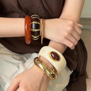 May include: A collection of five bracelets worn on the wrists. The upper wrist features a translucent orange bangle, a dark brown segmented bangle with gold accents, and a thin gold band. The lower wrist features a wide cream bangle with a central oval dark red stone in a gold setting and a smooth, polished gold-tone cuff.