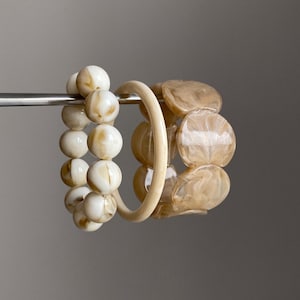 May include: A set of three bracelets featuring one strand of round, cream and tan marbled beads, one smooth, solid cream-coloured circular bangle, and one strand of flat, irregular, translucent tan and cream marbled disc-shaped beads.